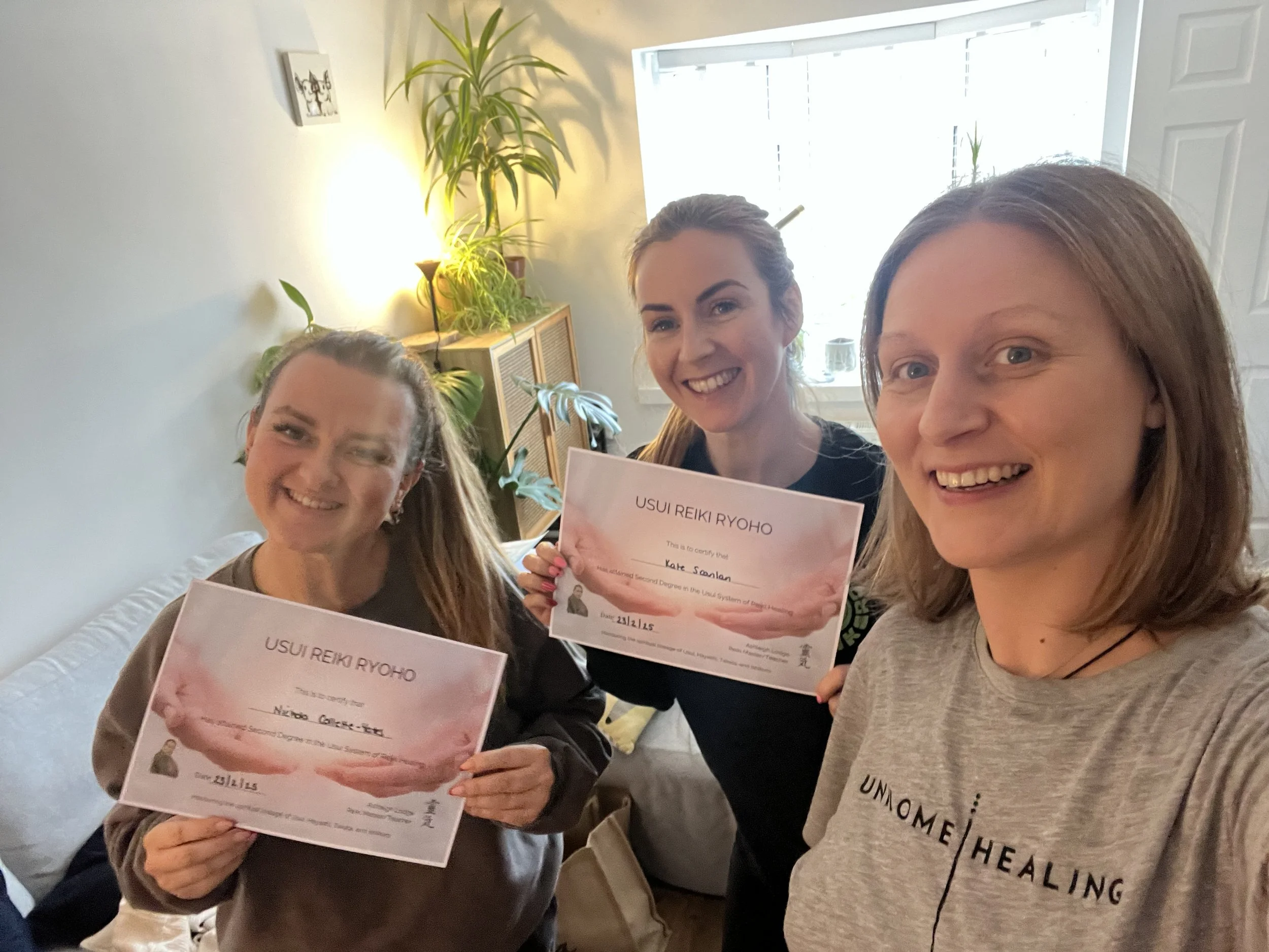 Three women smiling and taking a selfie indoors, two of them holding certificates for Usui Reiki Ryoho certification. The background features a window, some potted plants, and a wall with a small decorative picture.