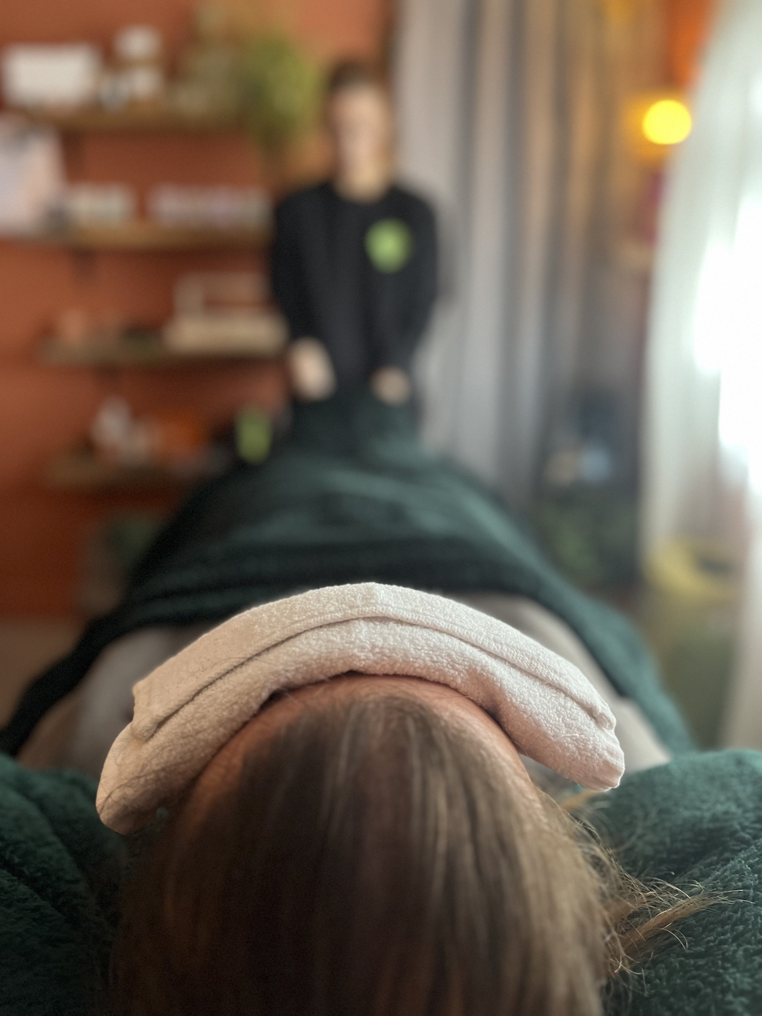 the top of a person's head as they lay on a therapy bed with a weighted eye pillow on and underneath blanket. The far end of the therapy bed is blurred and shows shelves, a person, and warm lighting in a cosy terracotta room