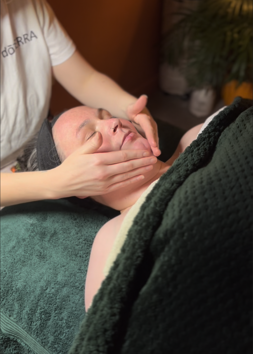 A woman receiving a facial massage while lying on a therapy bed with a green pillow under her head. A plush green blanket is covering their body, and a practitioner gently massaging their face.