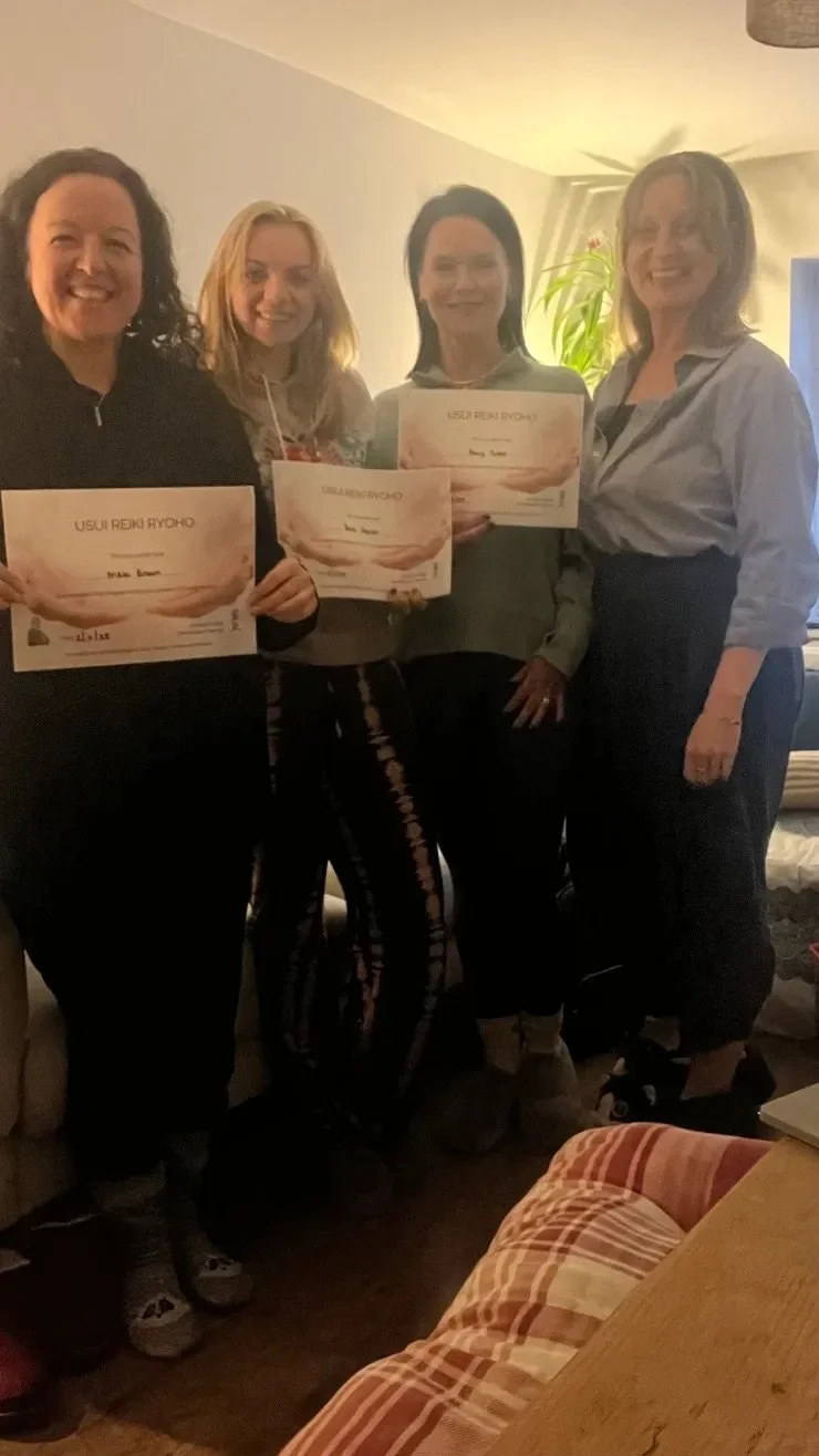 Four women smiling and holding Reiki certificates at an indoor event, standing in a living room with a white wall, a plant, and a table with a striped cloth.