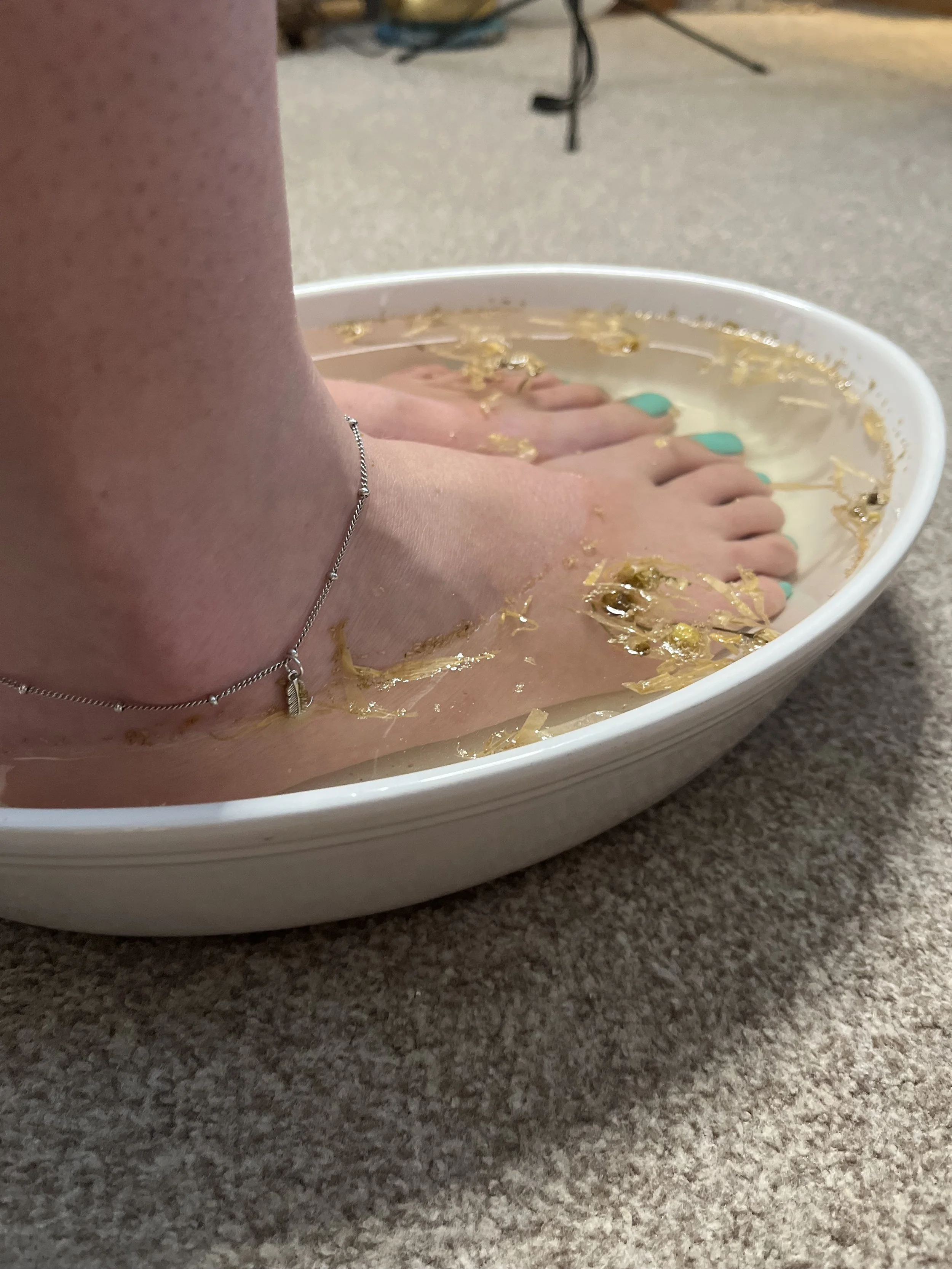 A person's foot with teal painted toenails soaking in a white basin of water with gold flakes.