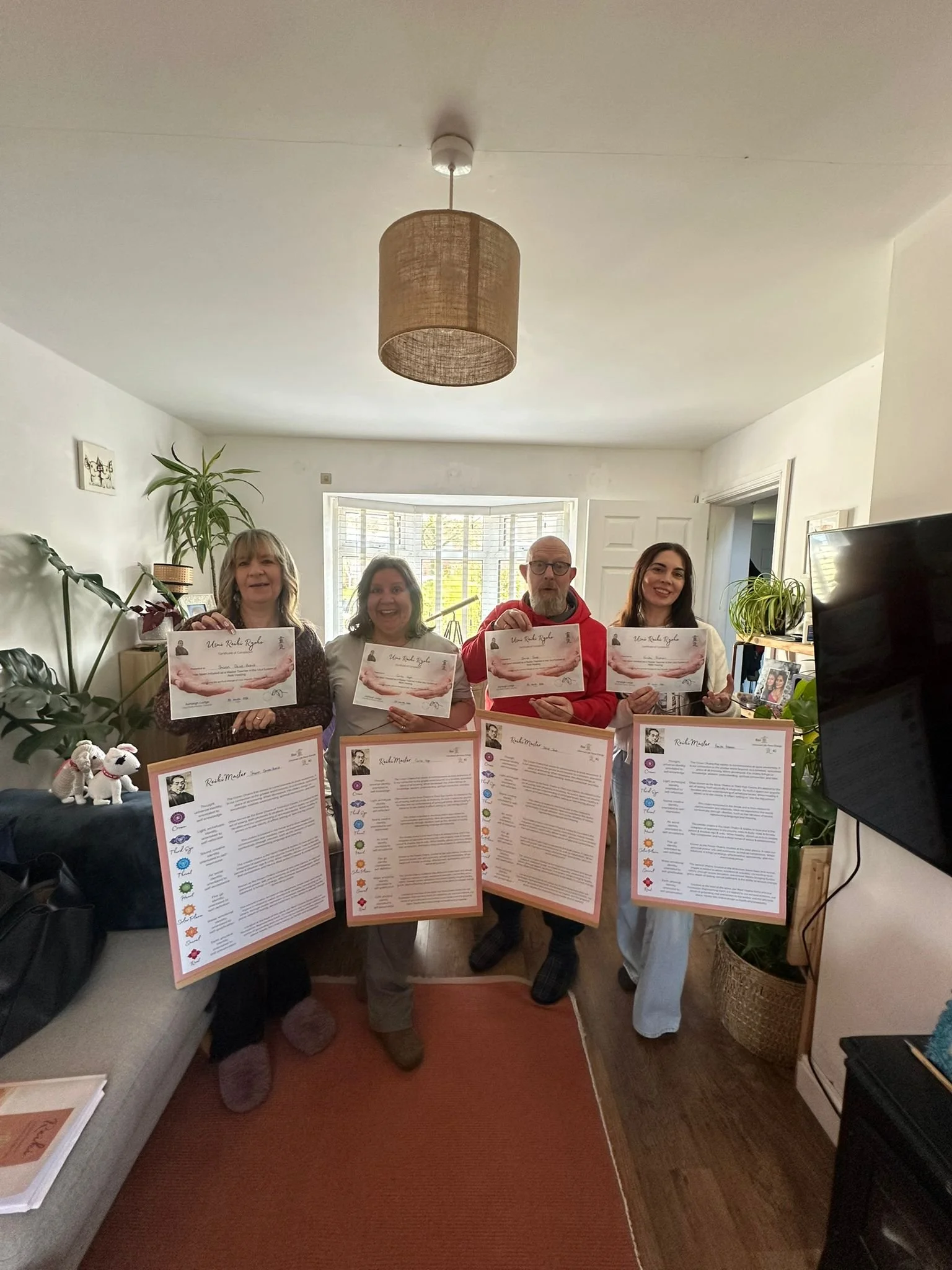 Four people standing indoors, holding certificates. The room has houseplants, a window with blinds, and a television. There are framed photographs and a small dog figurine on the table.