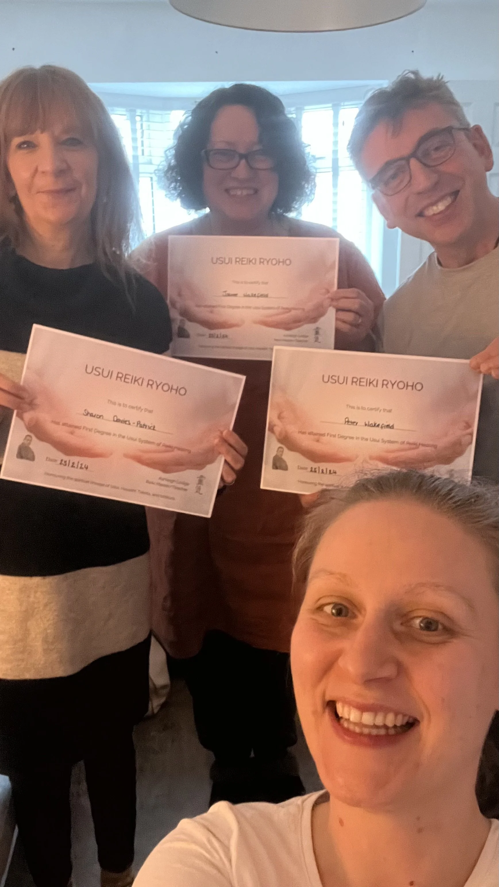 Three people smiling, holding Reiki certificates, with an extra woman at the bottom taking a selfie in a room with warm lighting.