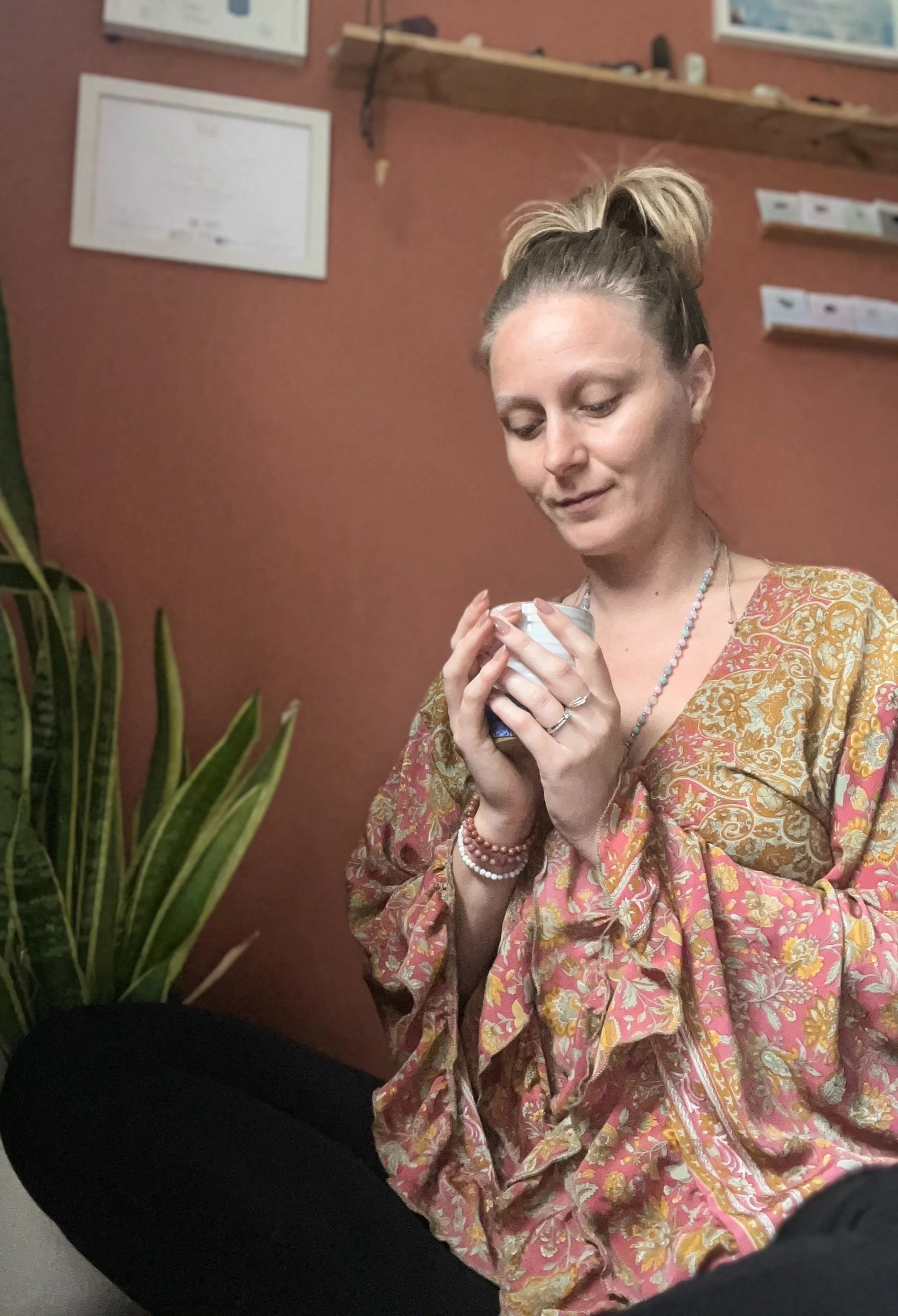 A woman sat drinking a cup of cacao, in a terracotta room looking contemplative and surrounded by plants, and certificates & crystals on the wall