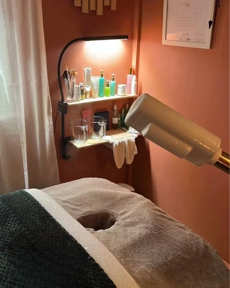 A warm inviting terracotta massage room with a treatment bed covered with a green blanket. There's a facial steamer and skincare products, and a light positioned at the side of the bed over some small shelves.