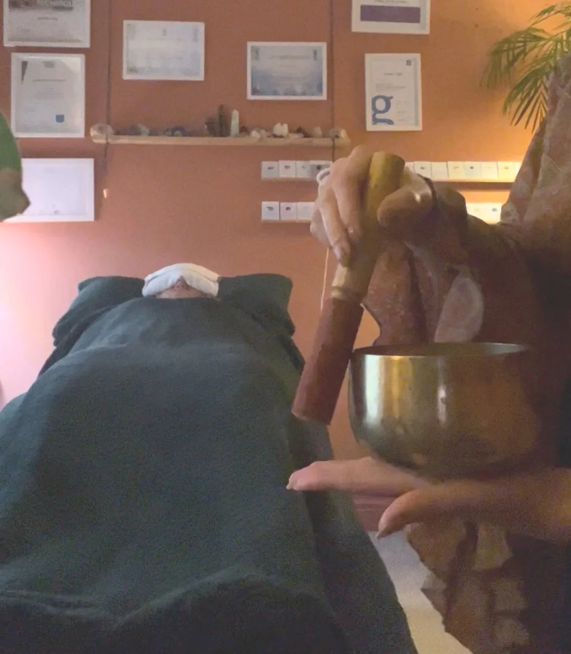 Person with weighted eye pillow lying on a therapy bed in a cosy terracotta room, with a therapist playing a large tibetan singing bowl around the bed.