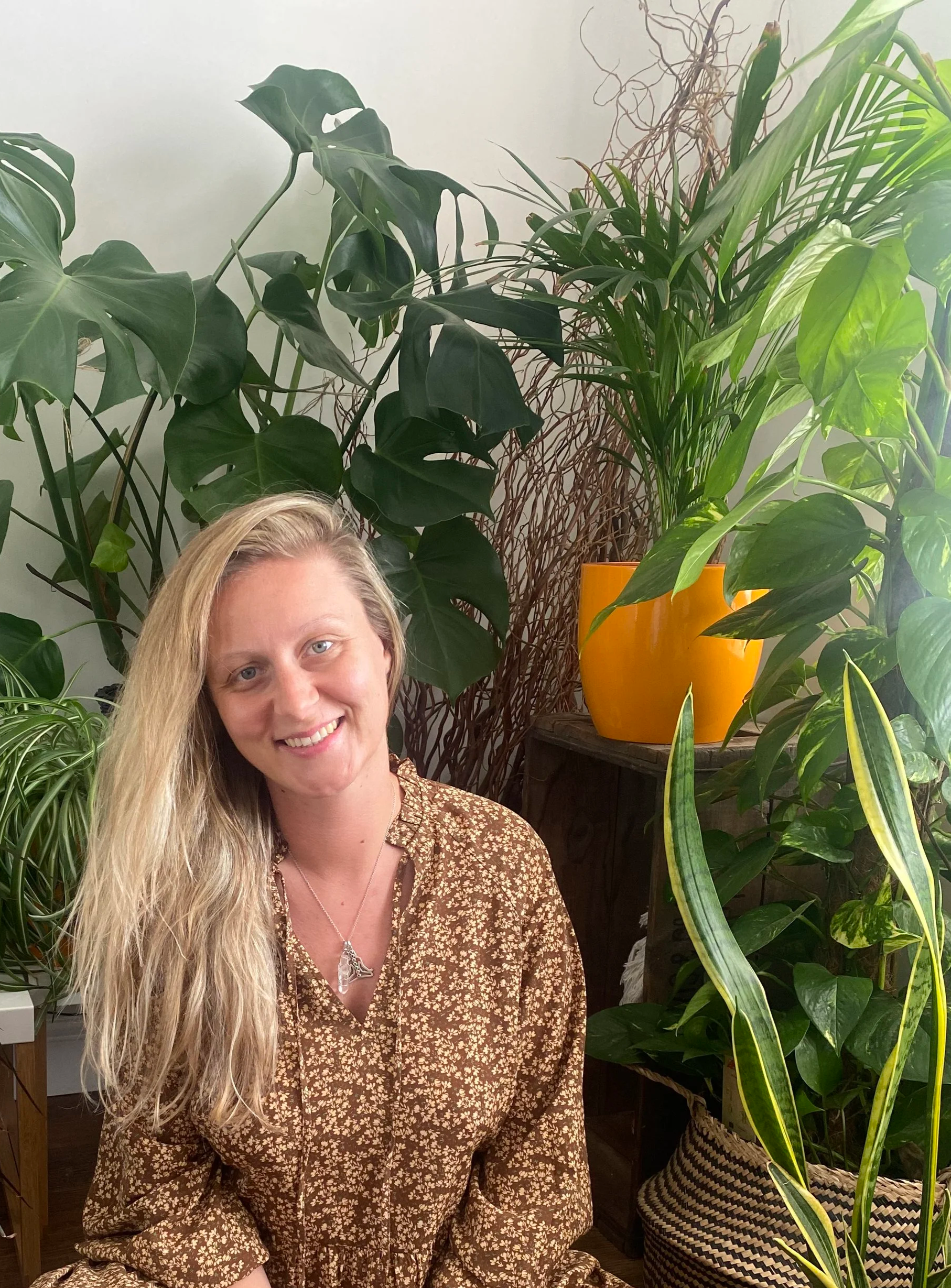 A woman with long blonde hair, blue eyes, and wearing a patterned brown dress with a silver necklace, is in front of a collection of large green indoor plants, including a Monstera and a palm, with a yellow pot in the background.