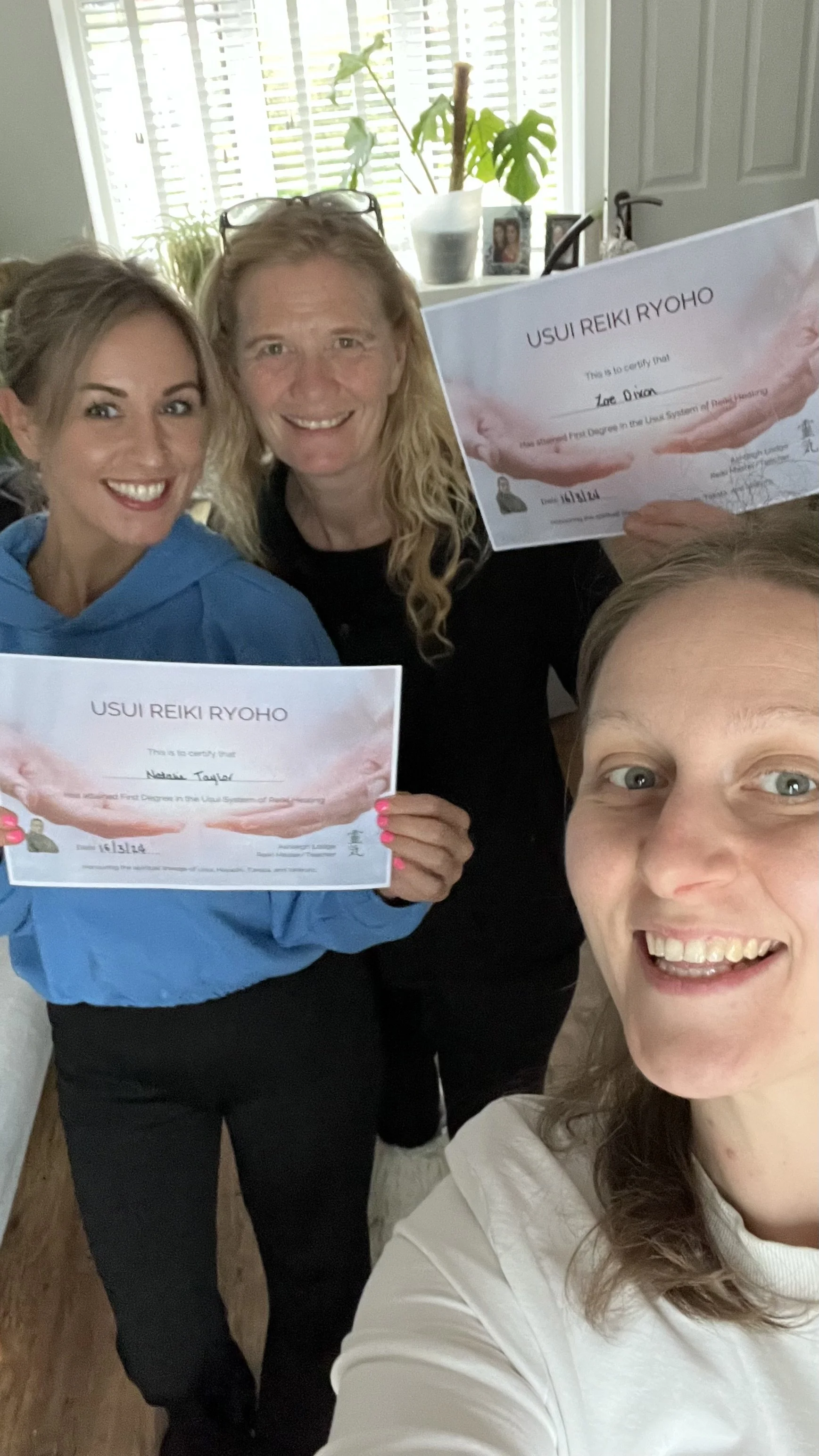 Three women taking a selfie together in a room. Two women are holding Reiki certificates, smiling. The background shows a window with blinds, some houseplants, and family photos.