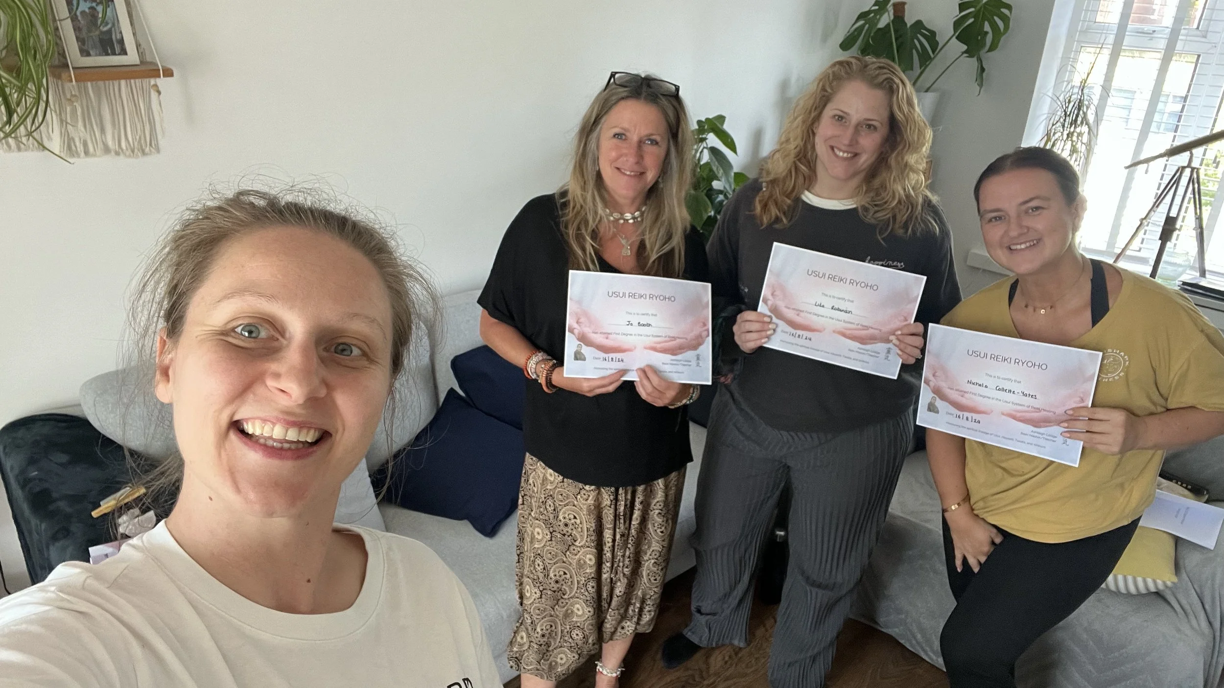 Four women posing together inside a room, three are holding Reiki certificates, with a plant, window, and decorative items in the background.