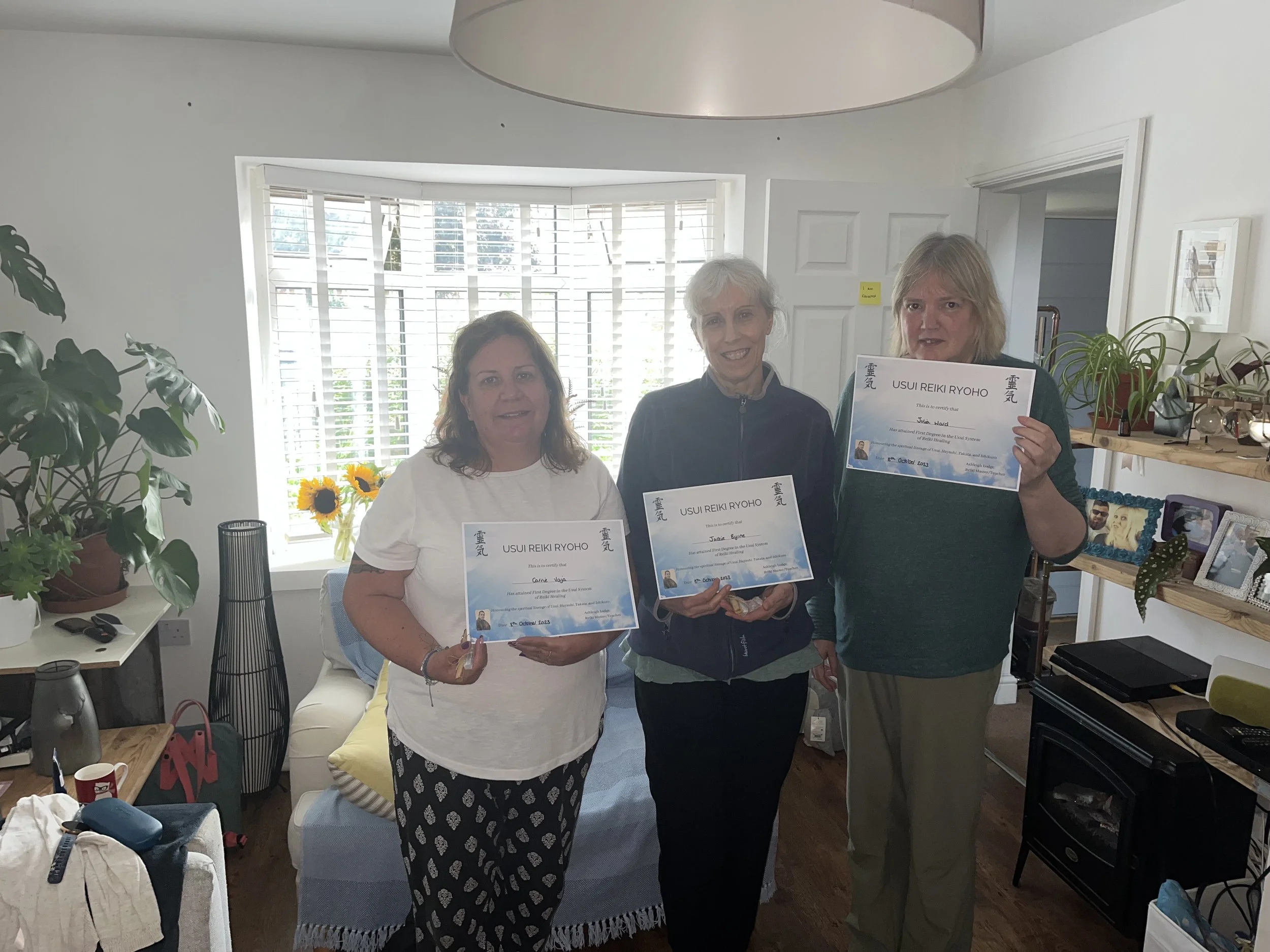 Three women standing in a living room, holding certificates for Reiki Ryoho, smiling at the camera. The room has a window with blinds, houseplants, and various decorations and personal items visible.