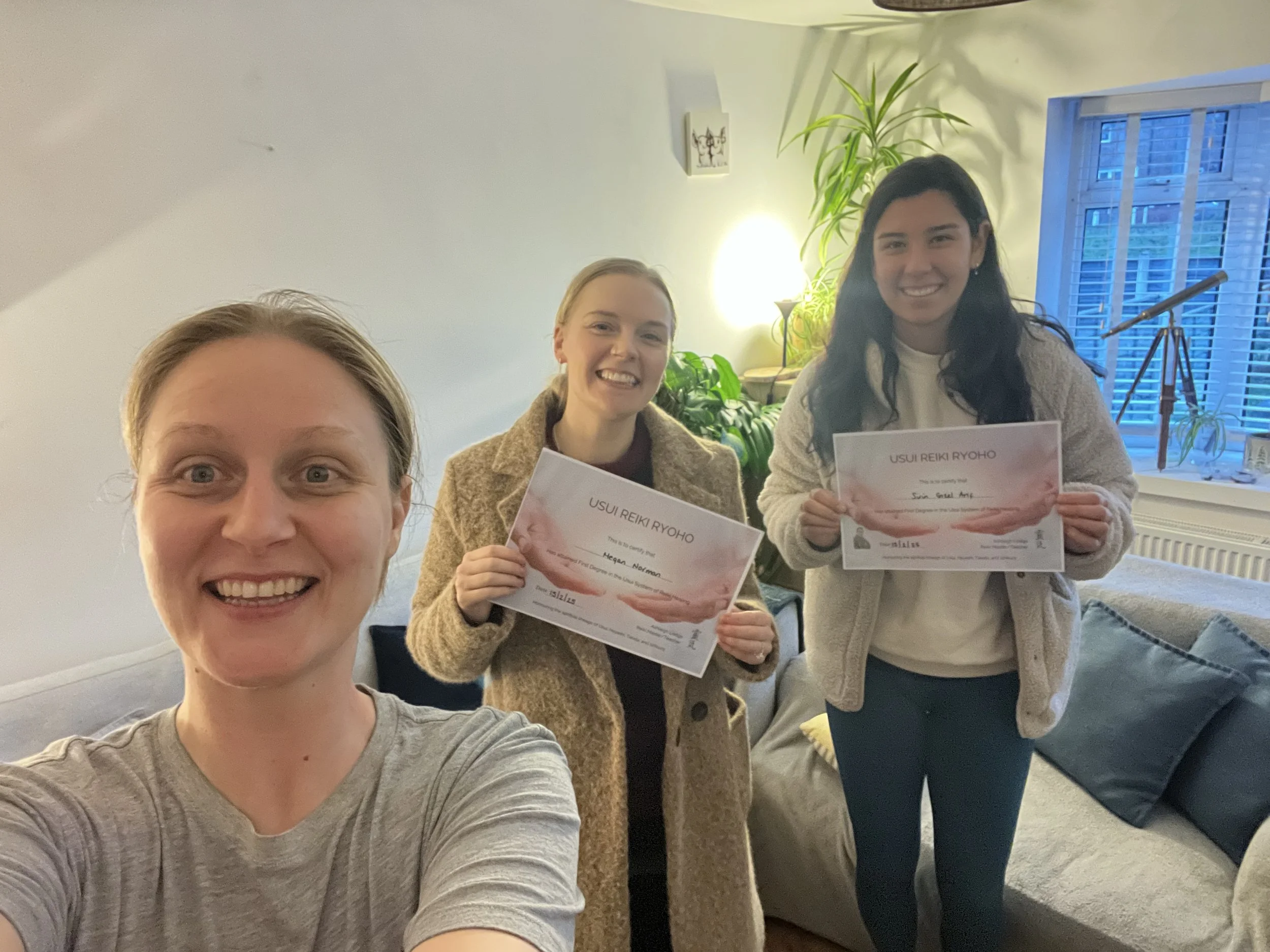Three smiling women, two holding certificates, standing in a cosy room with plants and a window with blinds.
