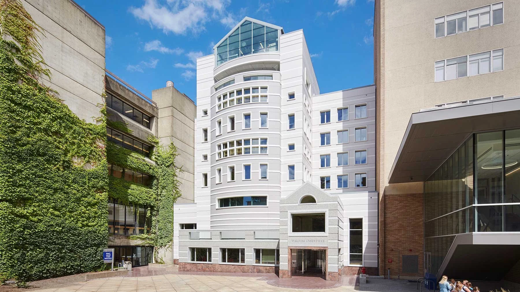 View of the Veilluem Institute building with modern white architecture, surrounded by other alley buildings, one with green ivy and large glass windows, under a blue sky with a few clouds.