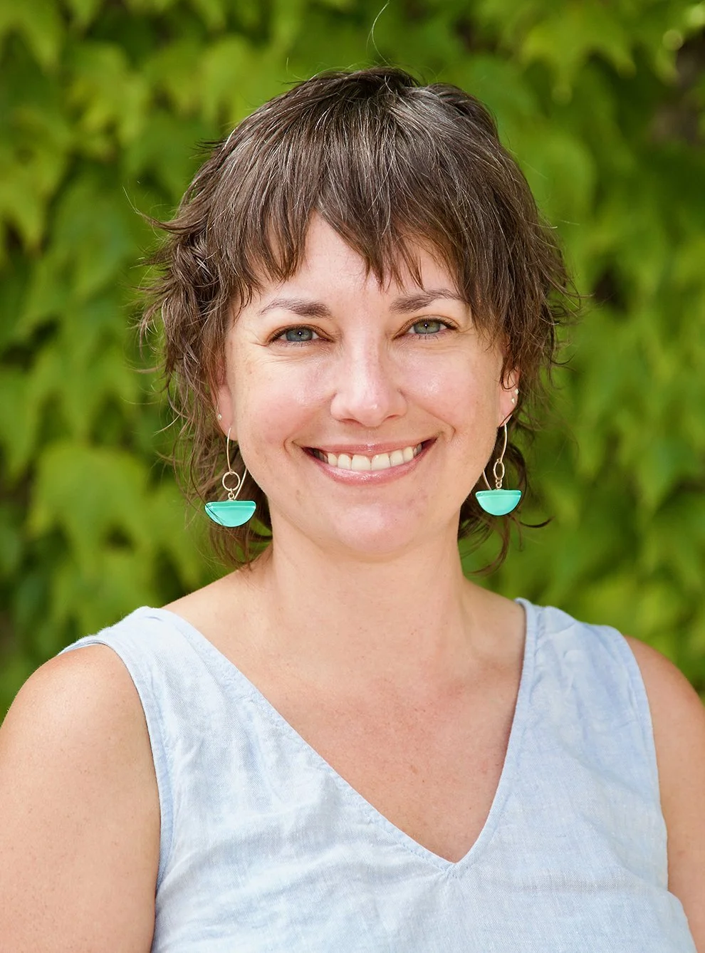 Smiling woman with short brown hair, wearing turquoise earrings and a sleeveless light blue top, standing outdoors with green leafy background.