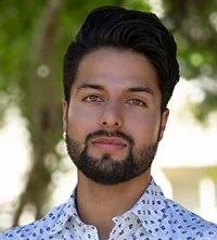 A young man with dark hair and a beard, wearing a light blue shirt with patterns, standing outdoors in front of trees.