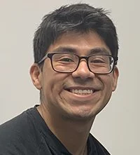 A smiling man with short black hair, glasses, and a black shirt standing against a plain background.