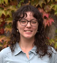 Young woman with curly dark hair and glasses smiling outdoors with autumn-colored leaves in the background.