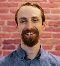 Portrait of a man with a beard and mustache, smiling, wearing a blue shirt, with a pink brick wall background.