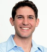 Headshot of a smiling young man with dark brown hair, wearing a light blue collared shirt.