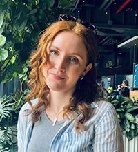 A woman with wavy red hair and blue eyes, wearing a light blue striped shirt, standing indoors with green plants and a dark background.