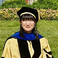 Woman in graduation cap and gown outdoors with green grass and yellow flowers in background.