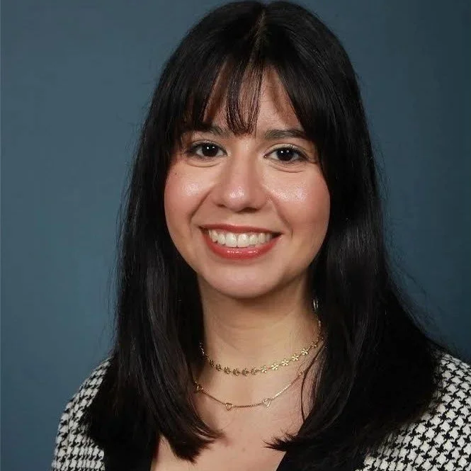 A woman with long black hair, bangs, and a bright smile, wearing layered necklaces and a black and white checkered top, standing against a blue background.