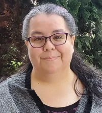 A woman with glasses and dark hair, wearing a gray sweater, standing outdoors with greenery in the background.