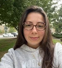 A young woman with long brown hair and glasses taking a selfie outdoors in a park, with trees and a few cars in the background.