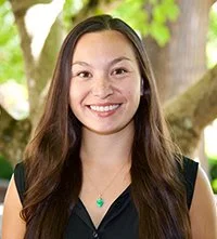 A smiling woman with long dark hair standing outdoors in front of green trees.