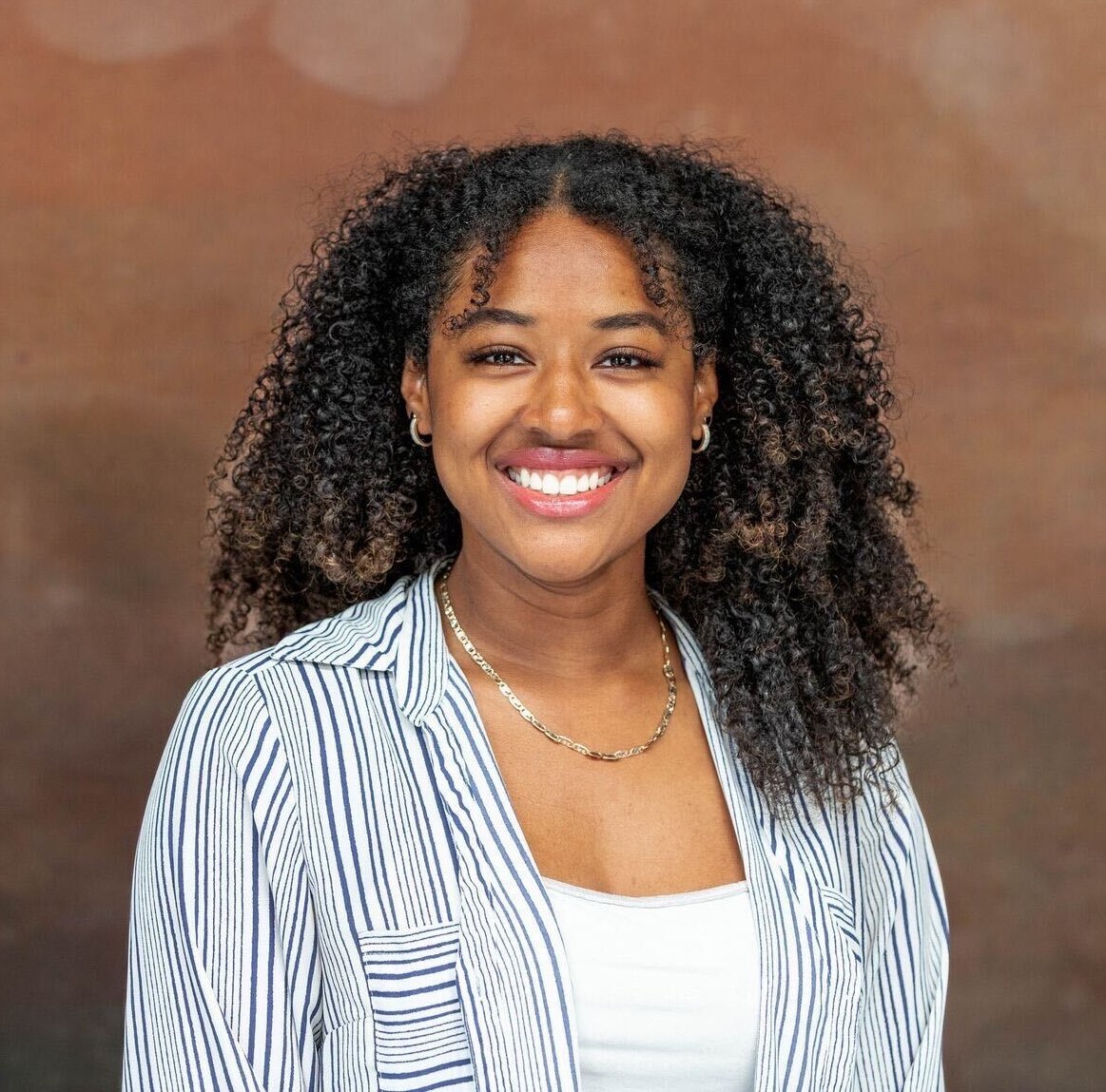 Portrait of a smiling young woman with curly hair, wearing a striped shirt, gold necklace, and hoop earrings, against a brown background.