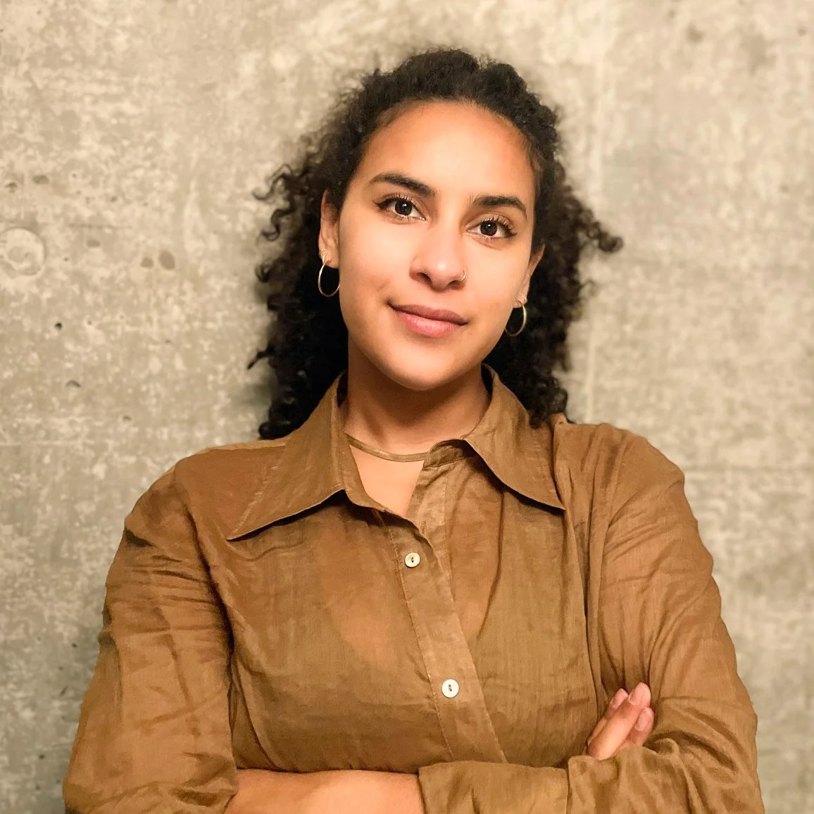 A young woman with curly dark hair, hoop earrings, and a nose ring, standing against a gray textured wall, wearing a brown button-up shirt with folded arms.