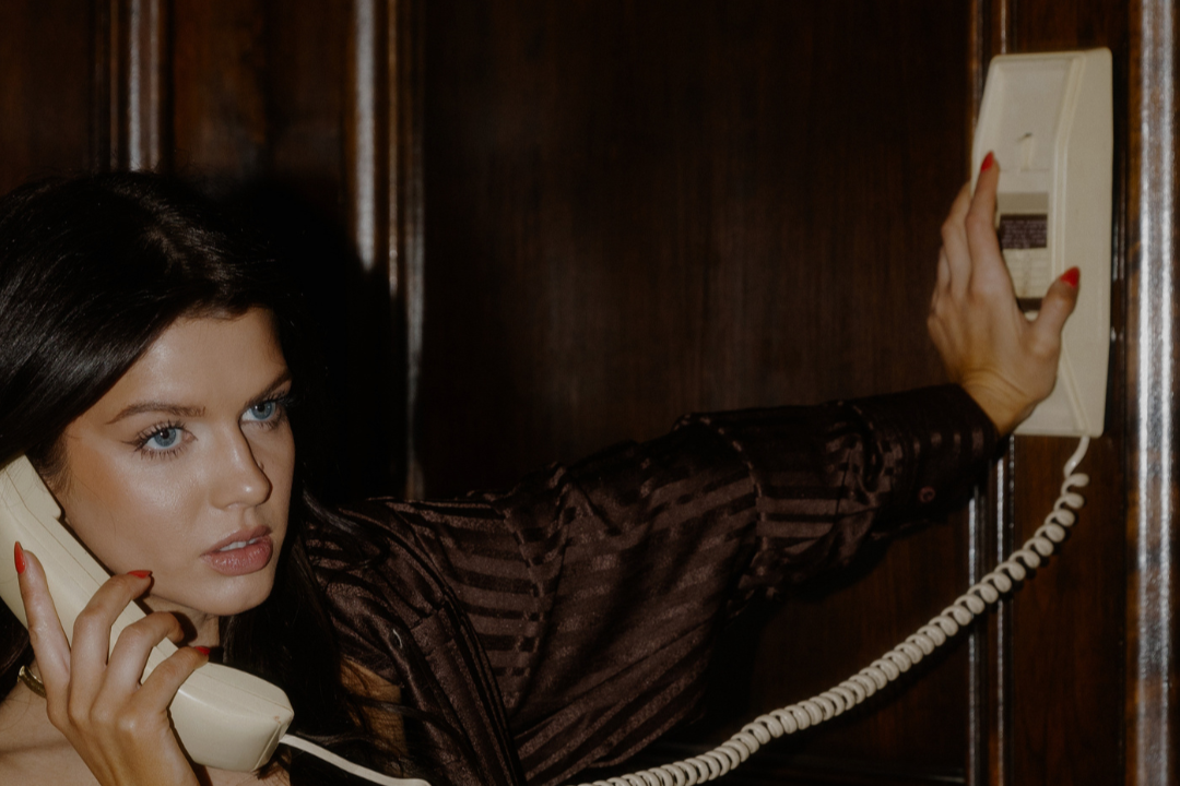 Woman with dark hair and blue eyes making a call on a beige landline phone, touching a wall phone with her other hand in a wooden-paneled room.