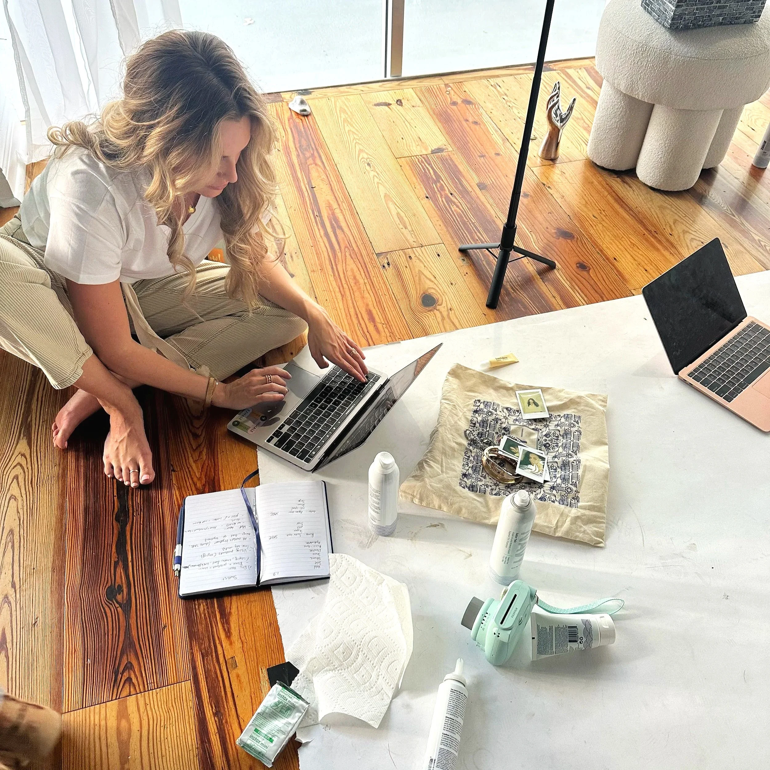 A woman with long curly hair sitting on the floor working on a laptop on a wooden table, surrounded by art supplies, printed photographs, and bottles of paint or glue. The scene appears to be in a bright room with large windows and curtains.