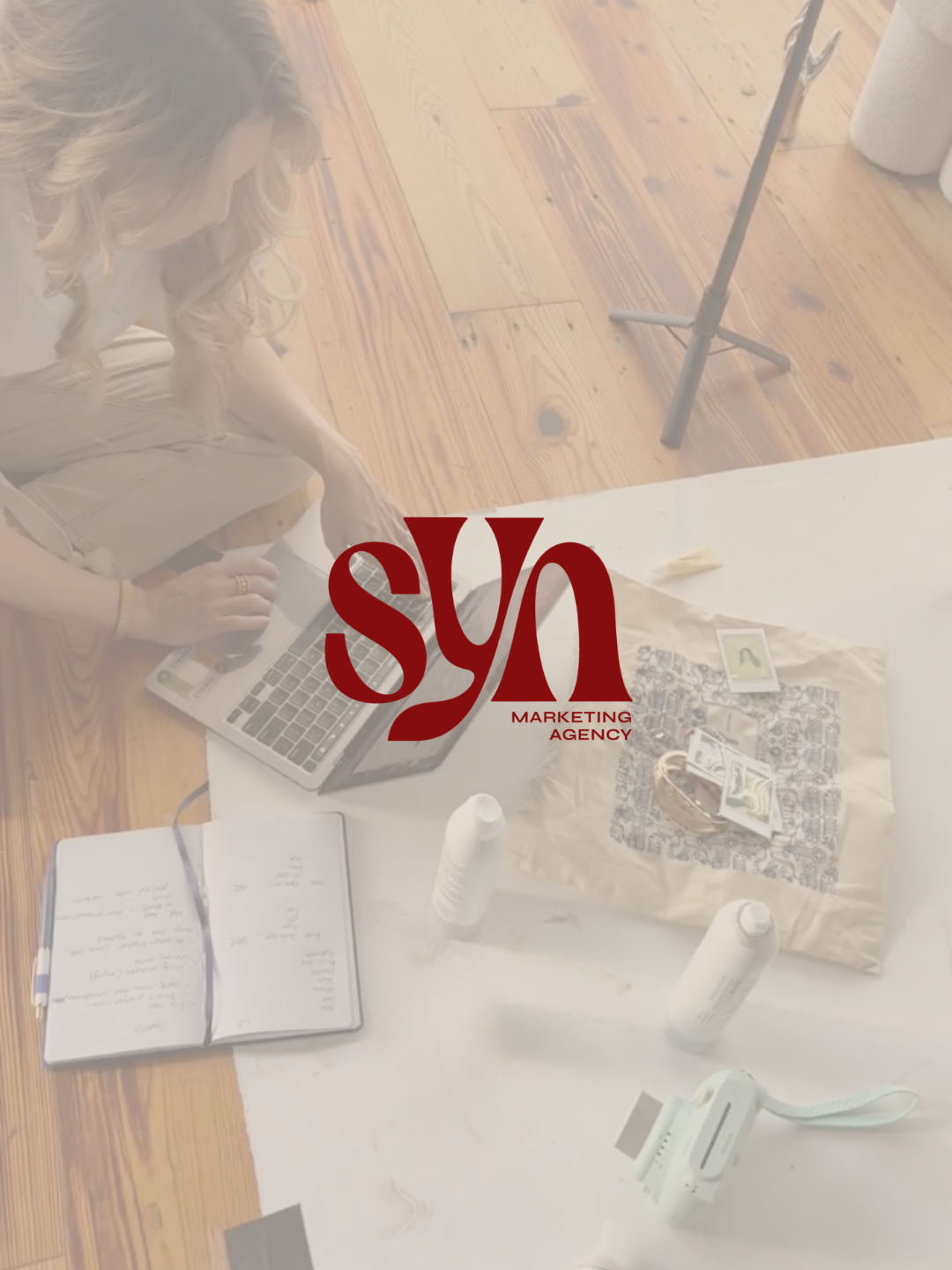 A woman with wavy hair and gold bracelet working on a laptop at a table with various items including a notebook, bottles, a small tray with pictures, and a camera. The photo has a semi-transparent red logo of a marketing agency.