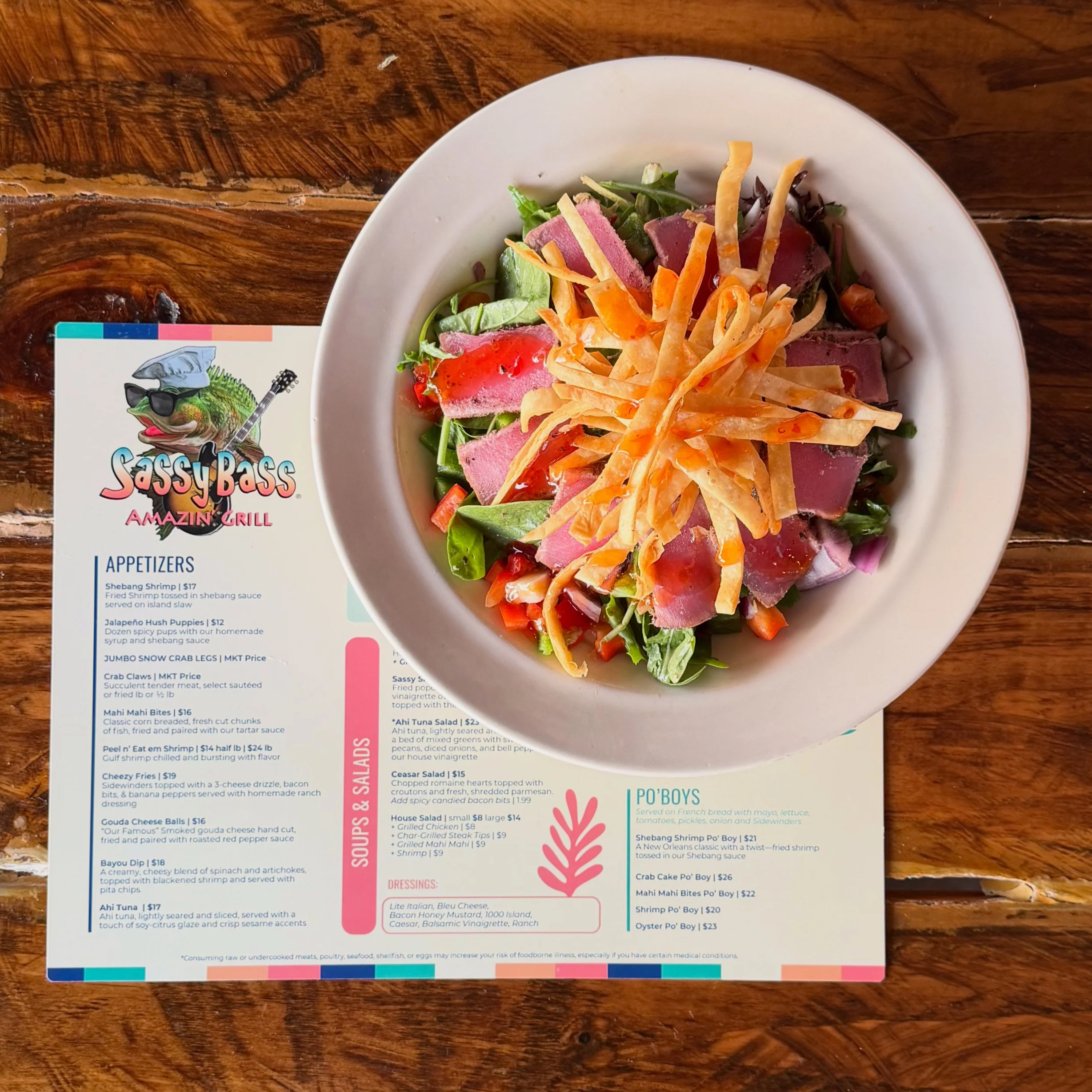 A white bowl containing a salad with greens, slices of roast meat, shredded cheese, and cherry tomatoes, on a wooden table. A menu and a colorful sticker featuring a fish with sunglasses and a guitar are partially visible near the bowl.
