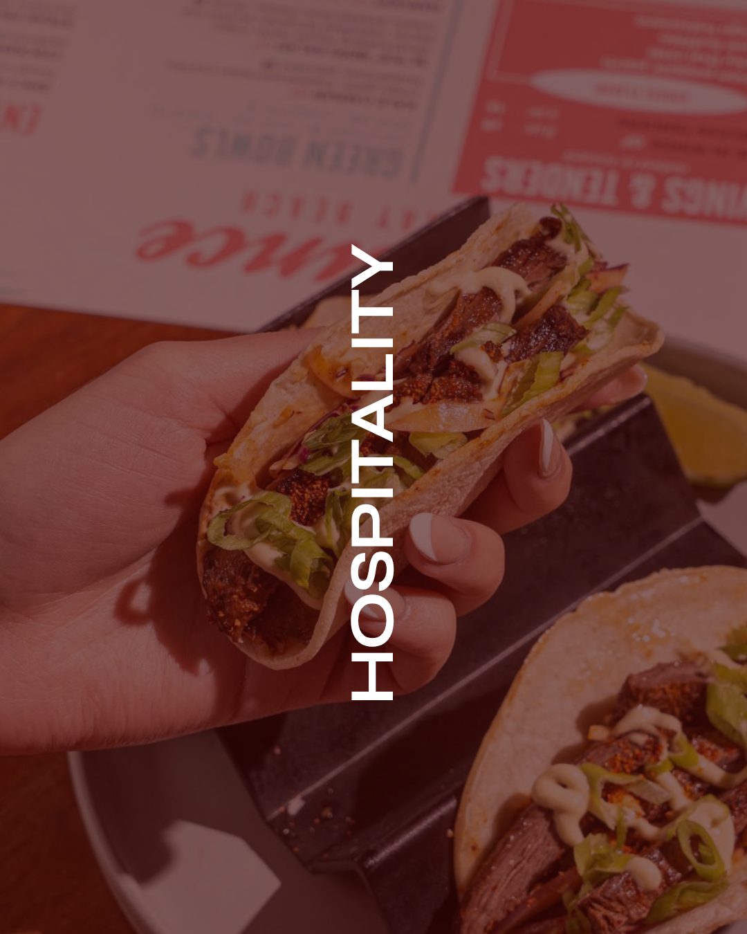 A hand holding a taco filled with grilled meat, lettuce, onions, and sauce, with a plate of similar tacos and a lemon wedge in the background.