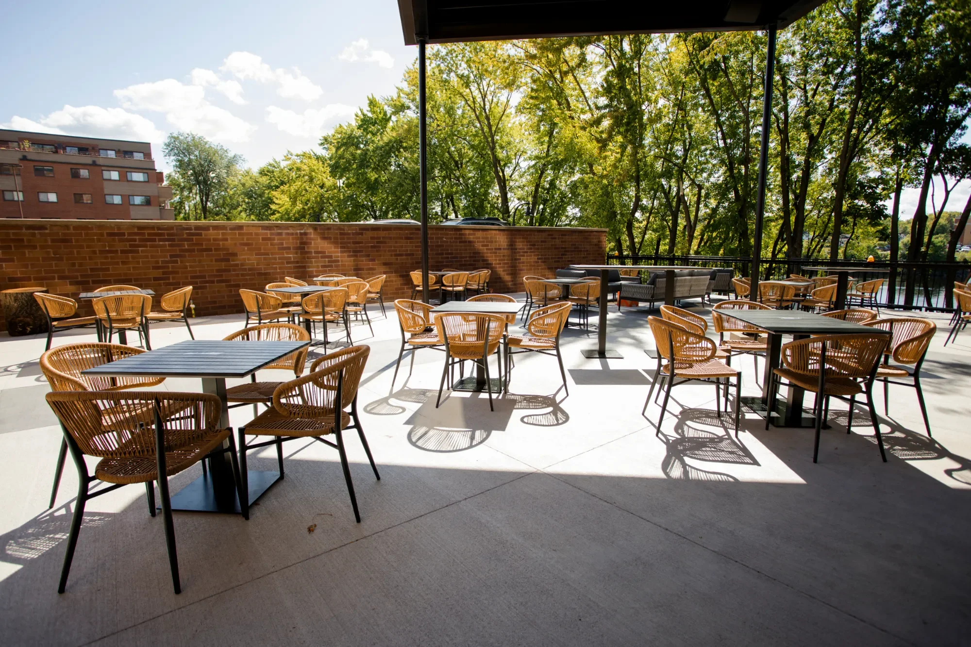 An outdoor patio with several round tables and woven chairs shaded by a roof, overlooking a green tree-lined area and a brick building in the background.
