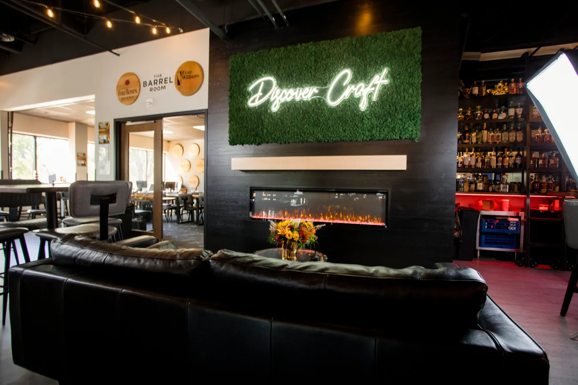 Modern lounge area with black leather sofa, a coffee table with a flower arrangement, a green wall with "Discover Craft" neon sign, a fireplace, and shelves with bottles of alcohol.