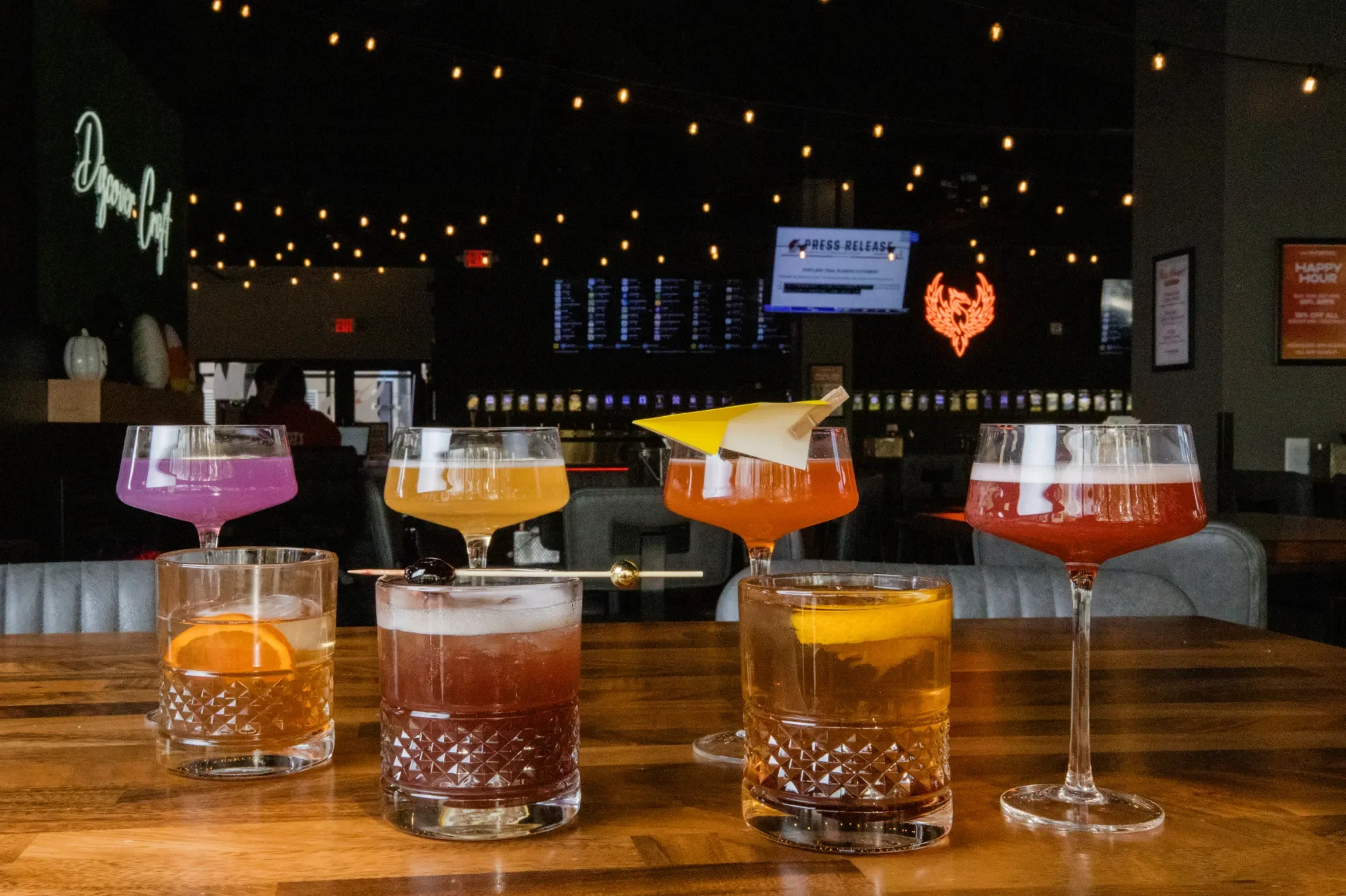 Multiple colorful cocktails with fruit slices on a wooden table in a dimly lit bar or restaurant.