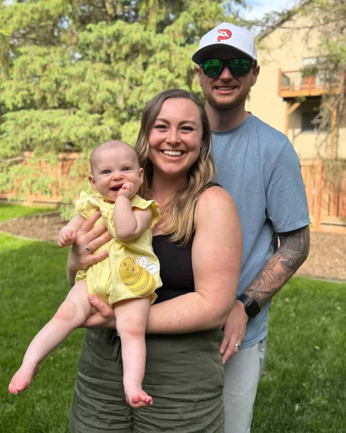 A happy family of three, a woman with long light brown hair, a man with a beard wearing sunglasses and a cap, and a smiling baby in a yellow dress, standing outdoors on a grassy lawn with trees and a house in the background.