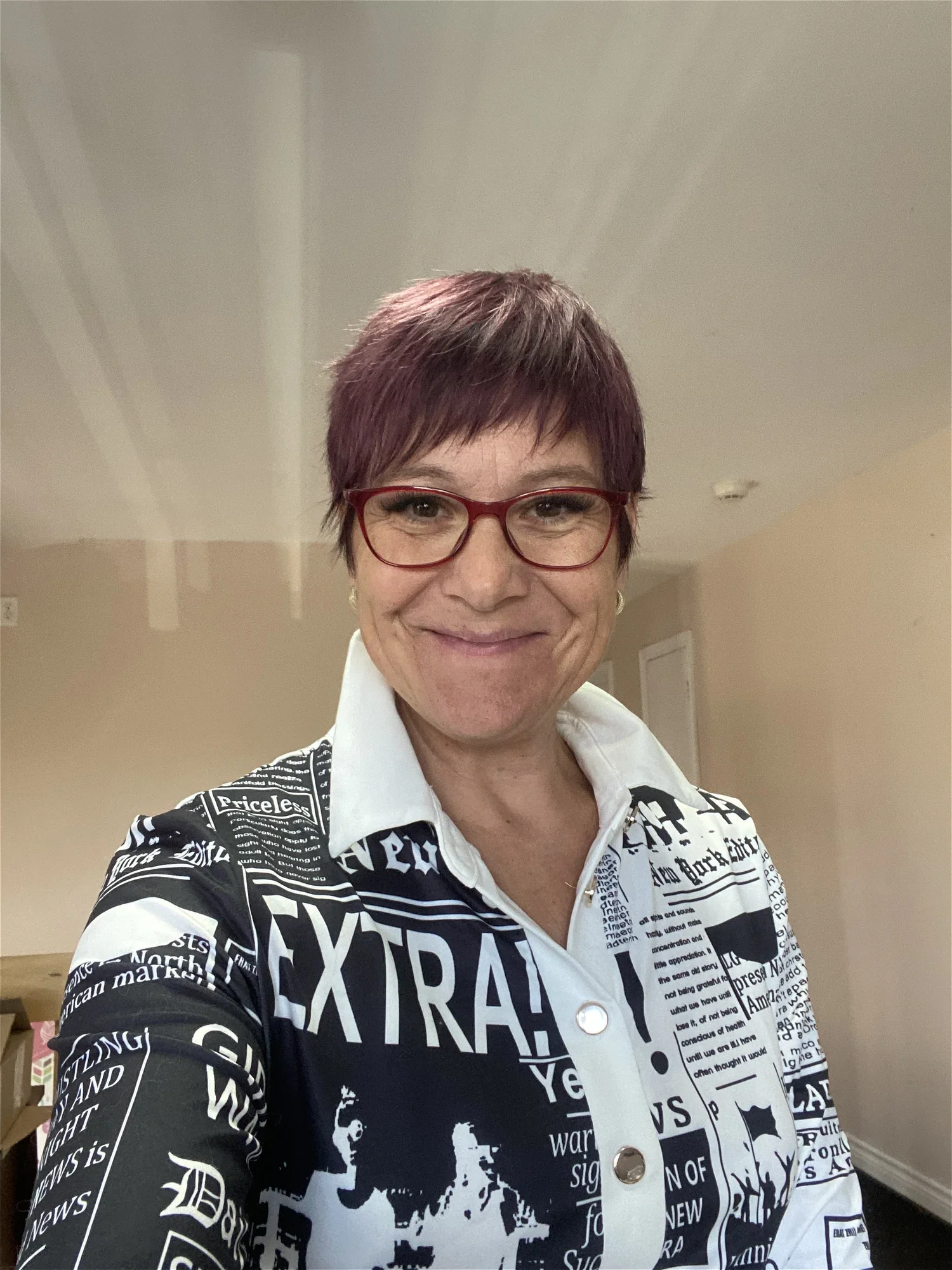 Une femme souriante porte des lunettes rouge et un manteau à motifs de journaux et de lettres, prenant un selfie dans un intérieur.