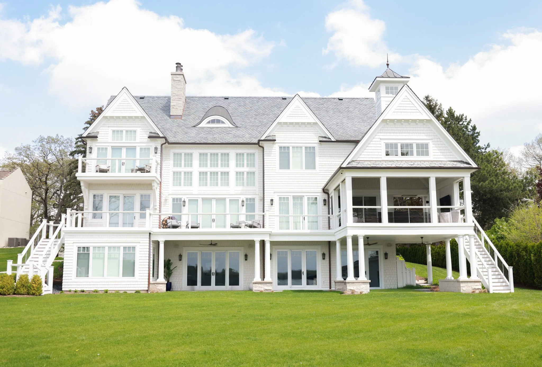 A large white house with multiple gabled roofs and a turret, surrounded by trees and a well-maintained green lawn, under a partly cloudy sky.