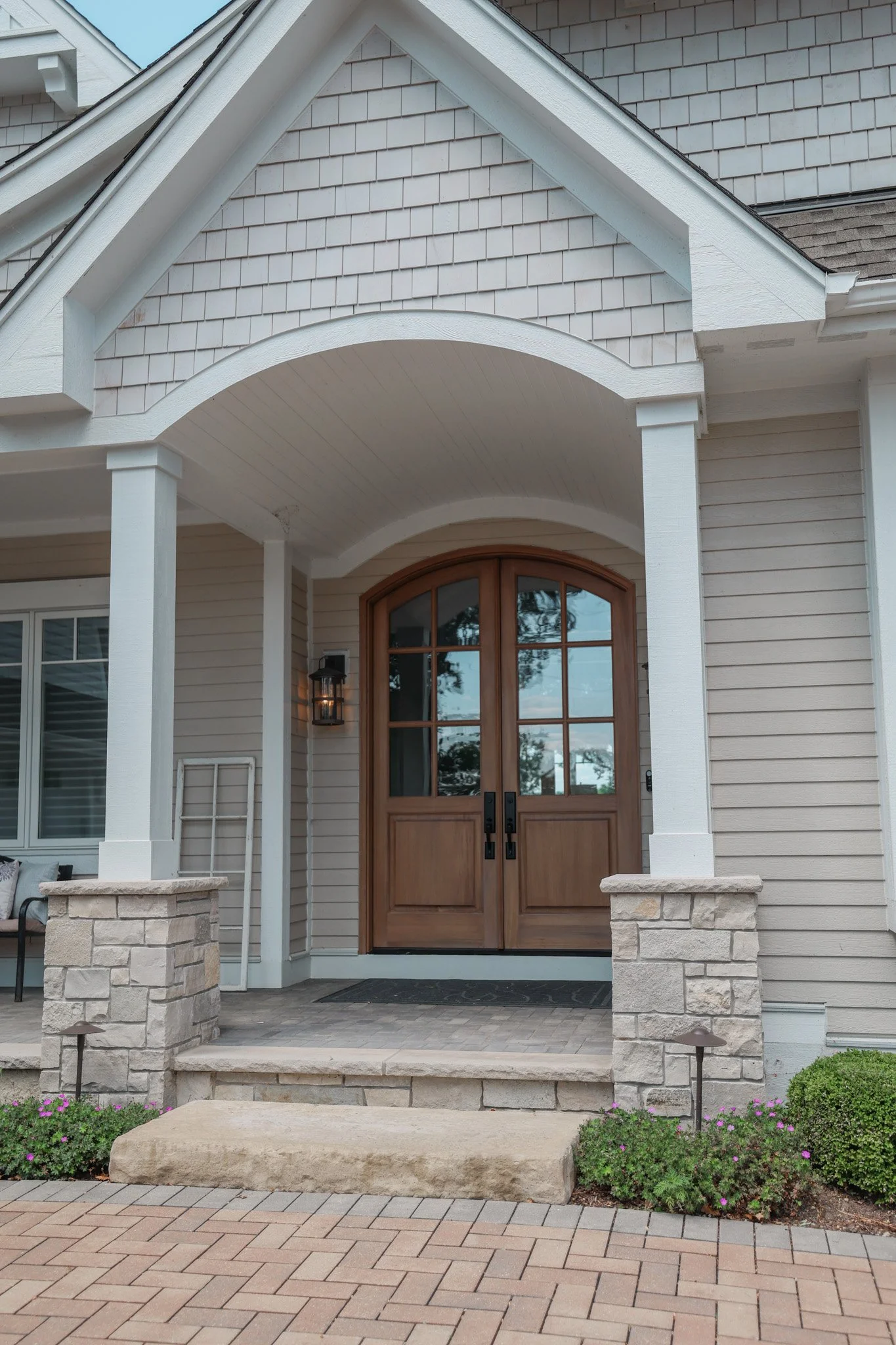 The house has a front porch with a wooden door, stone pillars, and a brick pathway leading up to it. Decorative plants are on each side of the path with purple flowers and small bushes.