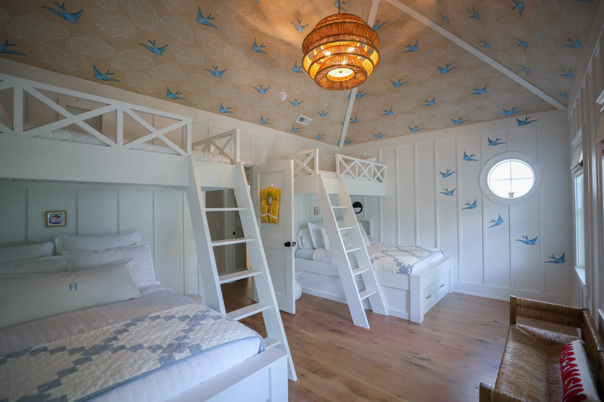 Bedroom with bunk beds, white panel walls, a circular window, and a ceiling decorated with blue birds and abstract clouds. There is a chandelier hanging from the ceiling, and bedding on the beds.
