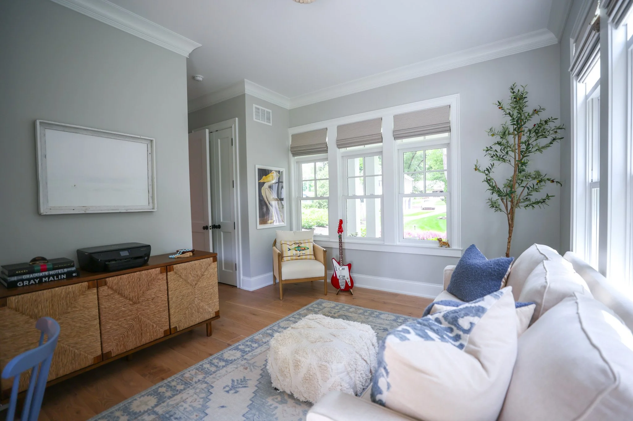 A cozy living room with large windows, a white sofa with blue and white pillows, a wooden side table with a printer and books, a small armchair with a pillow, a guitar, and a potted plant, with light-colored walls and hardwood floors.