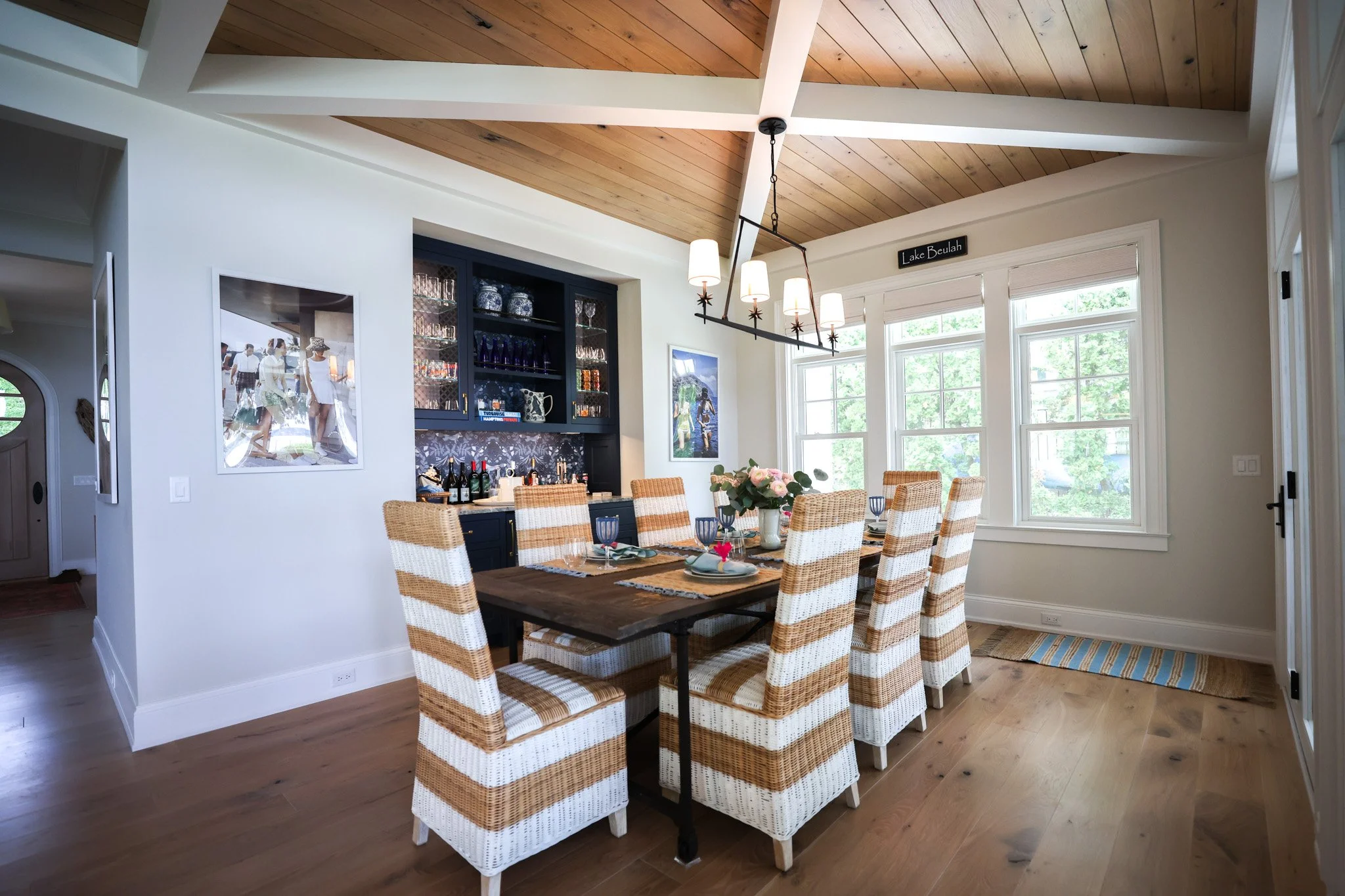 A dining room with a wooden table, seven wicker chairs with white and tan stripes, and a flower centerpiece. Large windows with white trim, a picture on the wall, a black cabinet with glass doors holding glassware and vases, a bar area with bottles, 
