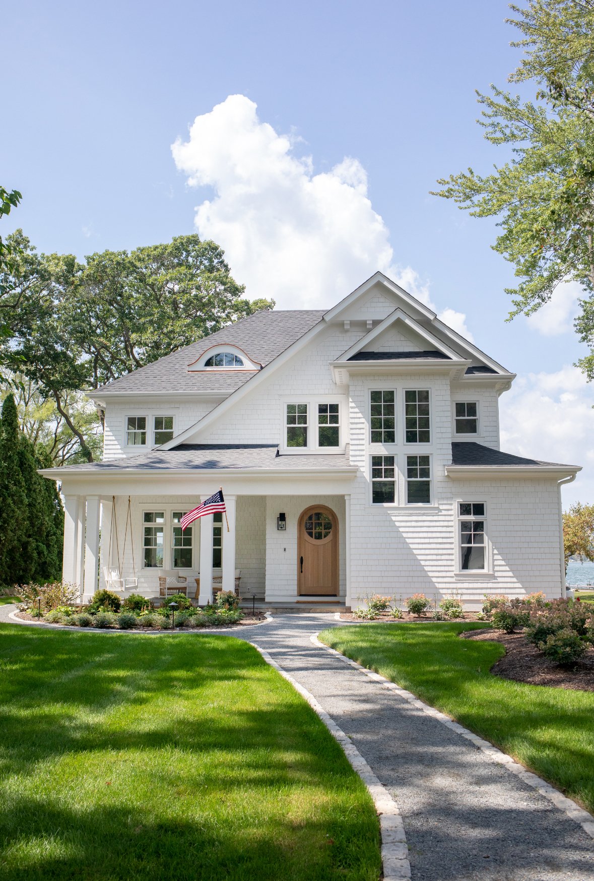 A white, two-story house with a manicured lawn, a curved pathway leading to the front door, and an American flag hanging near the porch. The house has multiple windows and a gabled roof, with trees in the background and a partly cloudy sky overhead.