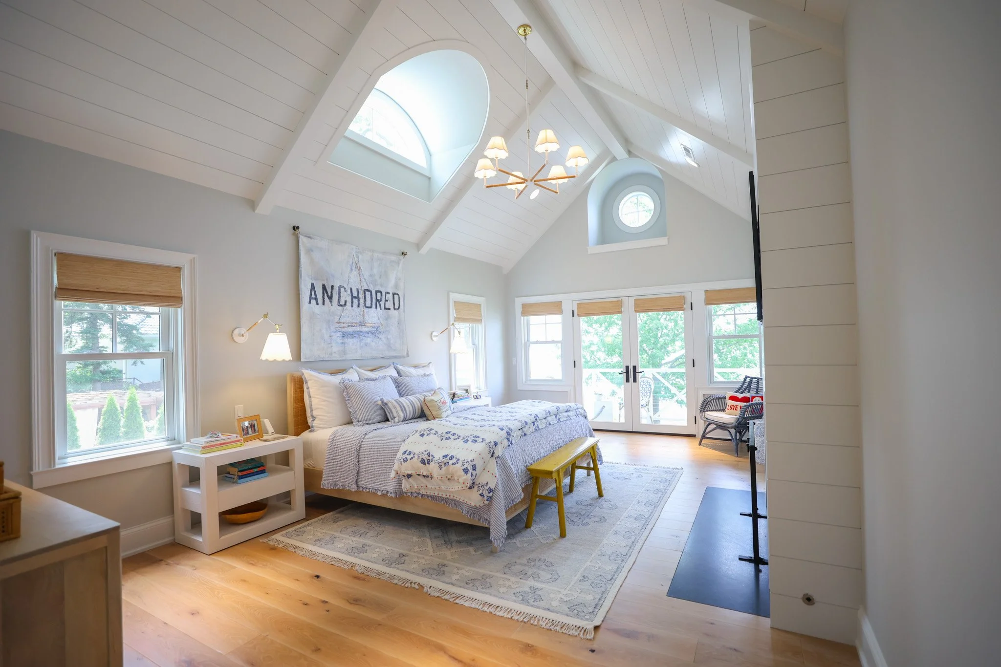 A bright bedroom with a vaulted ceiling, large windows, and a sitting area. A bed with white and blue bedding is near the windows, with a nightstand and framed photos. A chandelier hangs from the ceiling, and a blanket with the word 'ANCHORED' and a 