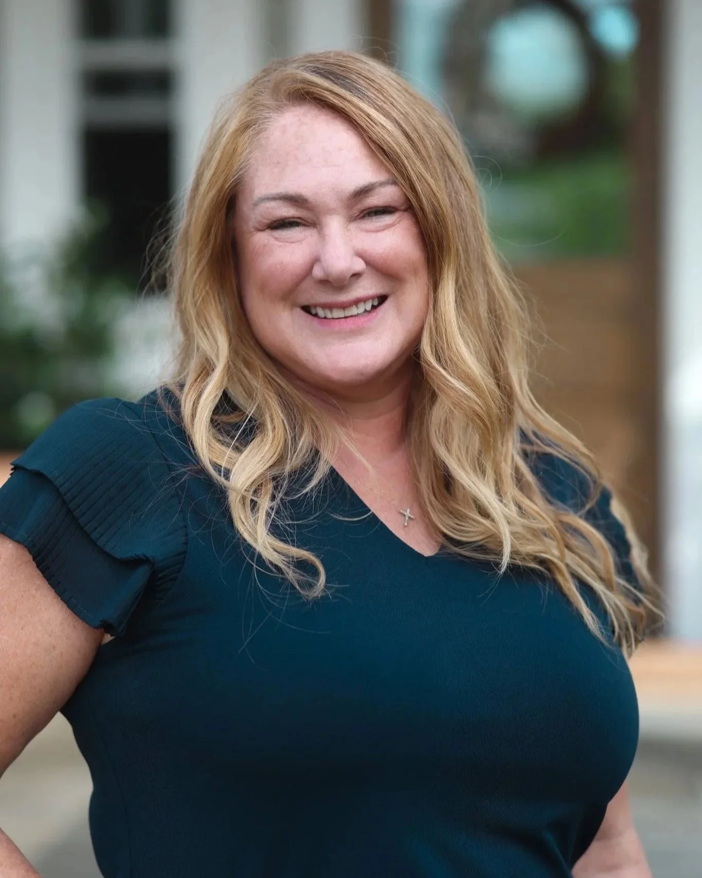 A woman with long, wavy blonde hair, smiling, wearing a navy blue top with ruffled sleeves and a cross necklace, standing outdoors with a blurred background of greenery and house.