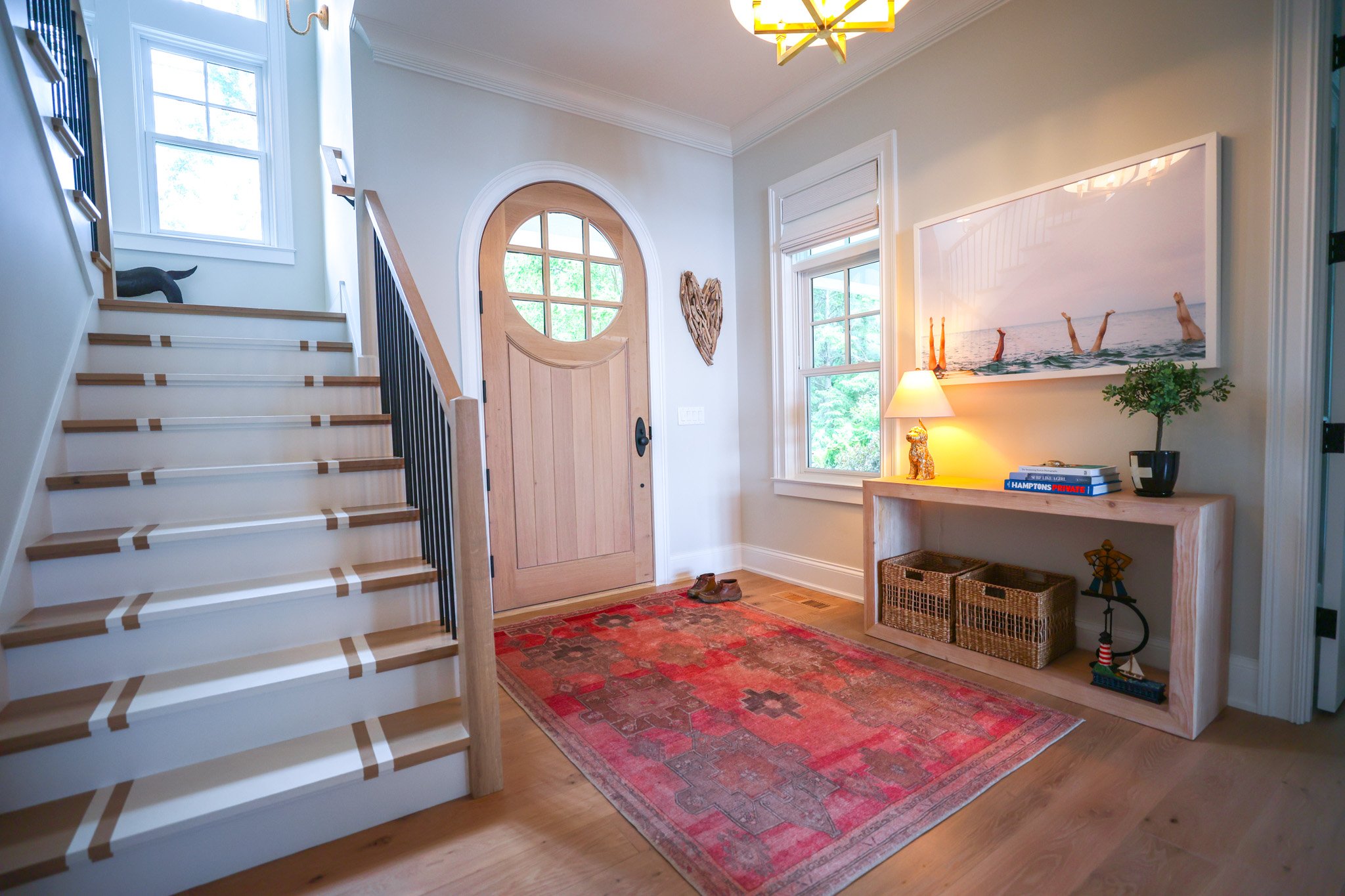 Entryway with staircase on the left, a front door with a circular window, a window on the right, a table with a lamp and decorative items, an artwork of feet in water hanging on the wall, a small potted plant, and a red patterned rug on hardwood floo