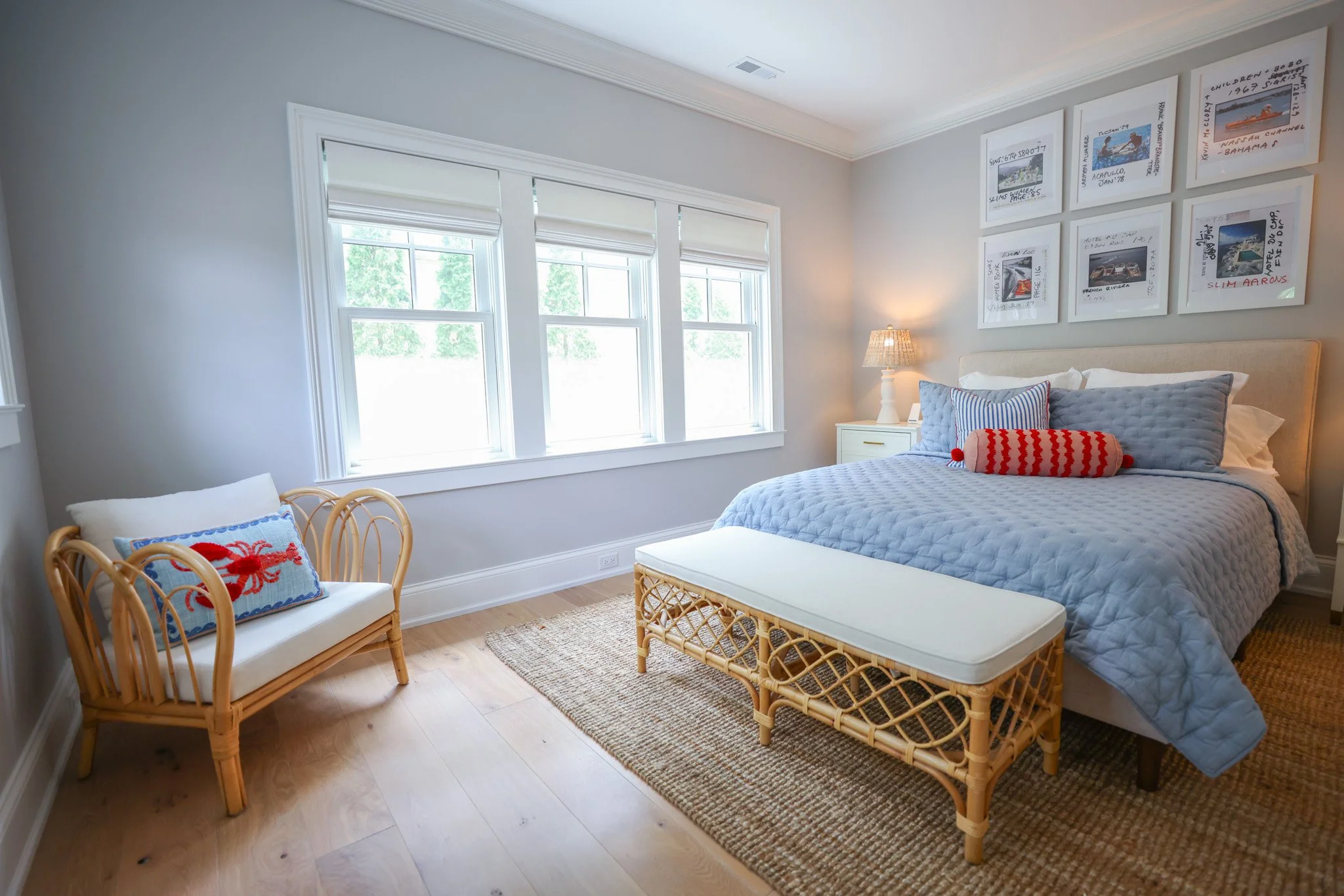 A cozy bedroom with a bed, a rattan bench, and two rattan chairs with cushions. The bed has a light blue quilt and pillows, and it is placed against a wall decorated with six framed pictures. There is a bedside table with a lamp and a window with whi